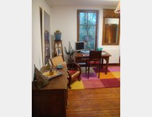 Ein gemütlicher Arbeitsraum mit Holzboden, einem Schreibtisch vor einem großen Fenster, einem Stuhl, einem Computer und einer Schreibtischlampe. Der Raum ist mit buntem Teppich, Pflanzen, einem Spiegel, einer Kommode und dekorativen Gegenständen eingerichtet.