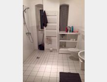 Modernes Badezimmer mit weißen Fliesen an Boden und Wänden, offener Duschbereich mit Duschkopf links, weißem Heizkörper mit Handtüchern in der Mitte und Waschbecken, Toilette sowie Regal mit Hygieneartikeln rechts. Ein kleines Fenster ist an der Rückwand sichtbar.