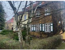 Zweistöckiges Wohnhaus mit gelber Fassade und roten Ziegeldächern, teilweise von kletternden Pflanzen bedeckt. Im Vordergrund stehen Birken und Büsche, die von einer gepflegten Gartenanlage umgeben sind.