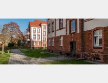 Backsteingebäude mit roten Ziegelfassaden und weißen Putzflächen im Stil einer historischen Wohn- oder Verwaltungsanlage, umgeben von einer grünen Rasenfläche und gepflasterten Wegen. Im Vordergrund eine Hauseingangstür mit Blumentöpfen, im Hintergrund weitere Gebäude und Bäume.