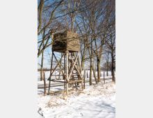 Ein hölzerner Hochsitz in winterlicher Landschaft, umgeben von schneebedecktem Boden und blattlosen Bäumen. Im Hintergrund ist ein Drahtzaun und eine weite, offene Fläche sichtbar.