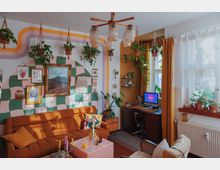 Wohnzimmer mit Retro-Dekor: Eine orangefarbene Couch steht vor einer Wand mit grün-rosa Schachbrettmuster, dekoriert mit Bildern und Pflanzen, die an Makramee-Seilen hängen. Rechts befindet sich ein Schreibtisch mit Computer vor einem Fenster mit gelben Vorhängen und zahlreichen Zimmerpflanzen auf der Fensterbank.