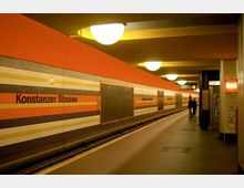 U-Bahn-Station "Konstanzer Straße" mit orangefarbener Wandgestaltung und horizontal verlaufenden Streifen in verschiedenen Farben. Die Bahnsteigbeleuchtung besteht aus runden Deckenlampen, und der Bahnsteig ist leer bis auf zwei Personen am rechten Bildrand.