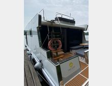 Hausboot mit moderner Ausstattung, das am Steg liegt. Sichtbar sind eine Außentreppe mit Rettungsring, Sitzbereiche mit Polstern auf der Terrasse und ein Schild mit der Aufschrift „The Kiss“.