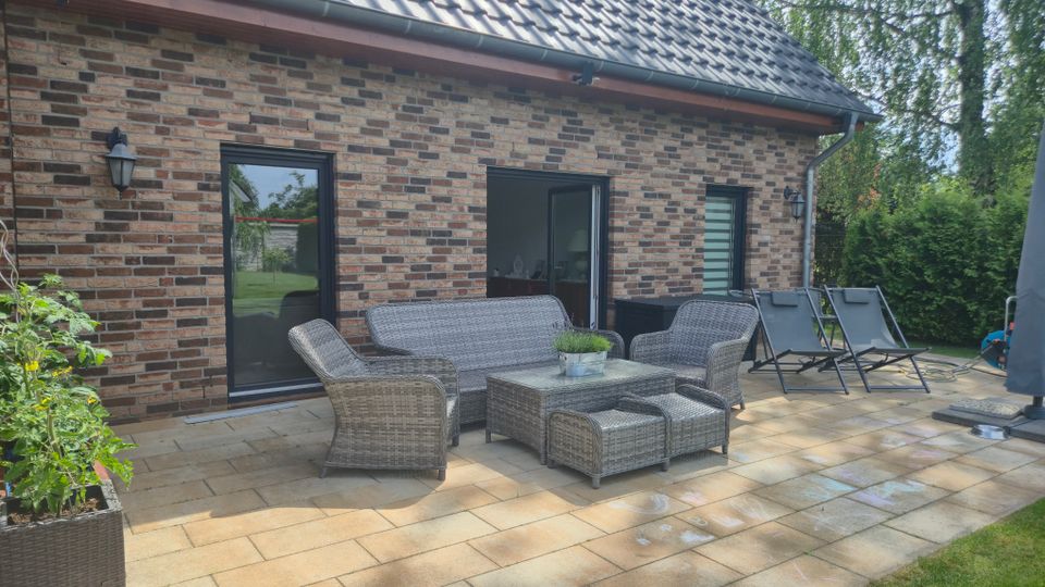 Gepflasterte Terrasse an einem Backsteinhaus mit Satteldach, Dachrinne und zwei Außentüren. Auf der Terrasse stehen eine Rattan-Sitzgruppe mit Tisch sowie mehrere Liegestühle; dahinter Gartenhecken und Bäume.