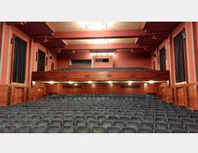 Innenraum eines Theatersaals mit vielen gepolsterten Sitzreihen, einer hölzernen Empore und roten Wänden mit dekorativen Elementen. Große Vorhänge und deckenmontierte Lichter sind sichtbar, der Raum wirkt symmetrisch gestaltet.