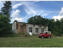 Ein verwittertes, einstöckiges Gebäude aus Ziegel- und Betonwänden mit teilweise beschädigten Fenstern, umgeben von Bäumen und Grasflächen. Im Vordergrund steht ein roter Geländewagen mit beigem Stoffverdeck.