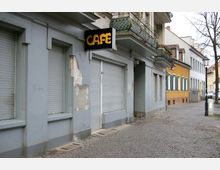 Straßenszene mit Gebäuden im Altbau-Stil, darunter ein geschlossenes Café mit grauen Rollläden und einem schwarz-gelben Leuchtschild. Der Gehweg ist mit Kopfsteinpflaster bedeckt, links sind Balkone mit dekorativen Gittern zu sehen, und rechts stehen Häuser mit farbigen Fassaden.