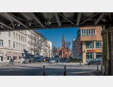 Straßenkreuzung in einem urbanen Viertel mit Blick auf eine rote Backsteinkirche im Hintergrund. Umgeben von alten, hellen Wohngebäuden und modernen Bauten; im Vordergrund eine Brückenkonstruktion und einzelne Fahrräder und Autos.