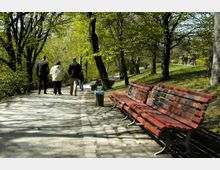 Ein Parkweg mit Kopfsteinpflaster, flankiert von Bäumen und grünem Gras. Auf der rechten Seite stehen rote, bunt bemalte Holzbänke, während mehrere Personen auf dem Weg spazieren.