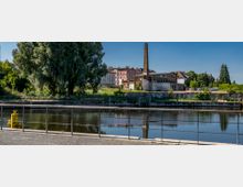 Industrielle Uferlandschaft mit einem Fluss im Vordergrund, umgeben von Geländern, und im Hintergrund eine verlassene Fabrik mit Schornstein, alten Backsteingebäuden und Bäumen. Wohnhäuser und Grünflächen ergänzen die Szenerie.