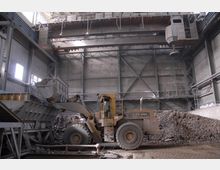 Industrielle Innenansicht mit einem Bagger, der Schutt in eine Metallschütte lädt. Der Raum ist von Stahlkonstruktionen, Betonwänden und einem hohen Deckenkran geprägt.