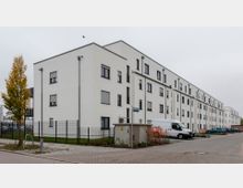 Mehrstöckiges modernes Wohngebäude mit weißer Fassade und zahlreichen Fenstern, entlang einer gepflasterten Straße. Im Vordergrund ein kleiner Verteilerkasten, ein geparkter Lieferwagen, sowie vereinzelte Autos und ein Baum mit herbstlichem Laub.