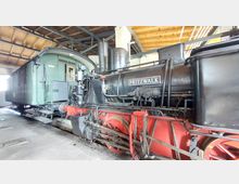 Historische Dampflokomotive mit der Aufschrift "Pritzwalk" steht in einer überdachten Halle. Im Hintergrund ist ein grüner Waggon erkennbar, und das Gebäude hat eine Holzdecke mit Stahlträgern.