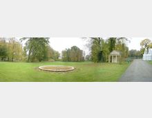 Weitläufiger Park mit einem runden Brunnen in der Mitte, umgeben von grüner Rasenfläche und großen Bäumen im Hintergrund. Rechts steht ein kleines pavillonartiges Gebäude mit Säulen, daneben ein Kiesweg.