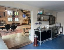 Moderner, offener Raum mit rustikaler Backsteinoptik. Im Vordergrund eine kleine Küchenzeile mit Metallarbeitsfläche, im Hintergrund ein Arbeitsbereich mit Tischen, Bürostühlen und künstlerischen Wanddekorationen; eine offene Treppe führt nach oben.