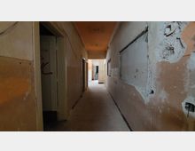 Ein leerer, heruntergekommener Flur in einem Gebäude mit sichtbaren Schäden an Wänden und Decke. Der Gang führt zu einem helleren Bereich mit einer Treppe im Hintergrund.