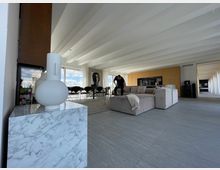 Weitläufiges modernes Wohnzimmer mit hellgrauen Fliesenboden, einer großen beigen Sofalandschaft, Holzverkleidung an der Wand und einer Glasfront, die viel Tageslicht hereinlässt. Im Vordergrund steht ein weißer Dekorationsgegenstand auf einem Tisch mit Marmormuster, im Hintergrund ist eine schwarze Sitzgruppe sowie ein großes Porträt an der Wand sichtbar.