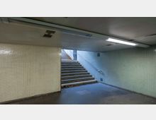 Unterführung mit gefliesten Wänden in hellen Farben sowie einer Treppe, die nach oben führt. Die Decke ist niedrig mit Leuchtstofflampen, und am Boden liegen einige Blätter.