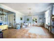 Eleganter Raum mit blauen Wänden, klassischen Möbeln im Rokoko-Stil und Kronleuchtern an der Decke. Der Raum ist mit Wandgemälden, Gemälden und einem Parkettboden gestaltet, durch eine Türöffnung ist ein weiteres lichtdurchflutetes Zimmer sichtbar.