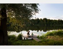 Ein kleiner Holzsteg am Ufer eines ruhigen Sees, umgeben von üppigem Gras und einem großen Baum im Vordergrund; im Hintergrund dichter Wald. Auf dem Steg sitzen ein erwachsener Mann und ein Kind, die auf das Wasser blicken.