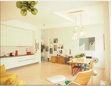 Offener Wohnraum mit integrierter Küchenzeile auf der linken Seite, ausgestattet mit weißen Schränken. Im rechten Bereich befindet sich ein Esstisch mit Stühlen, darüber hängen mehrere hängende Glühbirnenlampen. Die Wände sind mit Bildern und Dekorationen geschmückt, und der Raum ist hell mit viel Tageslicht.