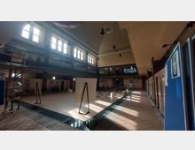 Innenraum eines historischen Schwimmbades mit hohen Decken, großen Fensterfronten und einem umlaufenden Balkon. Der Raum ist jetzt für Veranstaltungen umgestaltet, mit Sitzreihen, Informationstafeln und einer Bühne in der Mitte.