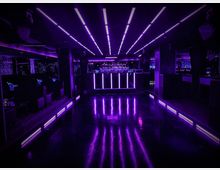 Ein Innenraum eines Nachtclubs mit dunklem Boden, lila Neonbeleuchtung an der Decke, den Wänden und entlang des Bodens. Im Hintergrund befindet sich eine beleuchtete Bar, umgeben von spiegelnden Oberflächen und Sitzbereichen.