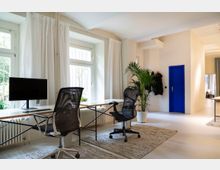 Helles, modernes Büro mit großen Fenstern, weißen Wänden und Vorhängen. Zwei schwarze Bürostühle stehen an einem Tisch, dahinter befinden sich Heizkörper, mehrere Pflanzen, sowie eine blaue Tür an der Wand.