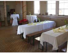 Ein rustikaler Veranstaltungsraum mit langer Festtafel und Bänken, die mit weißen Tischdecken und gelben Servietten dekoriert sind. Im Hintergrund stehen runde Stehtische, sowie alte, leicht verwitterte Wände und Fenster, die viel Tageslicht hereinlassen.