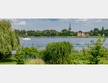 Ein Fluss mit grünem Ufer, im Vordergrund Schilf und eine Trauerweide. Im Hintergrund sind ein Segelboot, einige Häuser und eine Kirche mit spitzem Turm zu sehen.