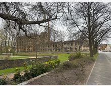 Historisches Gebäude mit einer Fassade aus Stein, umgeben von Bäumen ohne Laub, gelben Blumen und gemähtem Rasen sowie einem kleinen Fußballtor; im Vordergrund ein Gehweg mit einer Baumreihe auf der rechten Seite.