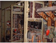 Detailaufnahme eines alten Schaltschrankes mit sichtbaren elektrischen Bauteilen wie Drähten, Spulen, Sicherungen und Anschlussklemmen. Der Schrankrahmen ist aus Metall, und die Kabel sind unorganisiert, teilweise verstaubt und abgenutzt.