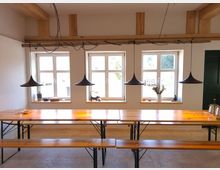 Ein heller Raum mit drei Fenstern, einem langen Holztisch mit passenden Bänken und vier schwarzen Hängeleuchten. Die Wände sind weiß gestrichen, die Decke hat sichtbare Holzbalken, und auf den Fensterbänken stehen verschiedene Gegenstände, darunter eine Vase mit Blumen.