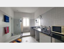 Helles Badezimmer mit rechteckigen, weißen Fliesen an den Wänden und auf dem Boden. Links ist eine ebenerdige Dusche mit Glasabtrennung, rechts ein schwarzer Waschtisch mit integriertem Waschbecken, Spiegel und verschiedenen Küchengeräten wie einer Mikrowelle. In der Mitte befindet sich ein Fenster mit Blick auf einen Heizkörper.