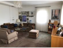 Wohnzimmer mit beigefarbenem Teppichboden, einer Sitzecke aus braunen Polstermöbeln, einem runden Glastisch in der Mitte und Bilderrahmen an der weißen Wand. Ein großes Fenster mit transparentem Vorhang lässt Tageslicht herein, daneben stehen ein Bücherregal und eine Holzkommode mit Dekorationsgegenständen und Fotos.