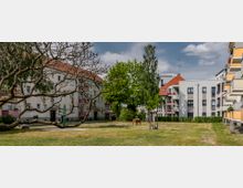 Wohnanlage mit mehrstöckigen Gebäuden, umgeben von Rasenflächen und Bäumen. Im Vordergrund ein baumloser Ast und eine Holzbank, im Hintergrund Gebäude mit roten Ziegeldächern und Balkonen.