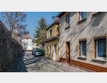 Schmale, gepflasterte Gasse mit einem gelben Wohnhaus rechts und einer Steinmauer links, im Hintergrund ein Fachwerkhaus mit rotem Dach. Parkende Autos entlang der Straße und Bäume im Hintergrund unter klarem Himmel.