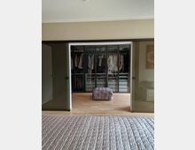 Schlafzimmer mit Blick auf einen begehbaren Kleiderschrank, der durch Glasschiebetüren abgetrennt ist. Im Vordergrund befindet sich ein Bett mit einer gesteppter Bettdecke, in der Mitte ein gepolsterter Hocker auf einem Holzfußboden.