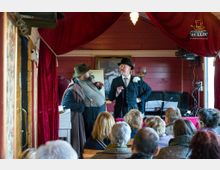 Eine kleine Bühne in einem rustikalen Raum mit roten Holzwänden und Vorhängen, auf der zwei Männer in historischer Kleidung ein Gespräch führen, während ein Publikum auf Holzbänken zuschaut. Im Hintergrund sind ein Gemälde, Notenständer und Beleuchtungselemente sichtbar.