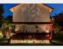 Eine Outdoor-Bar in einem Garten vor einer hellen Hauswand, beleuchtet mit warmen Lichtern. Die Bar ist dekoriert mit Pflanzen, Palmen in Töpfen und leuchtenden Elementen, umgeben von Rasen und Bäumen im Abendlicht.