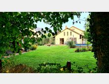 Ein gelbes Einfamilienhaus mit Satteldach, großen Fenstern und einer Terrasse, umgeben von einem gepflegten Garten mit Rasenflächen, Pflanzen und Bäumen. Im Vordergrund ein Baum und Blattwerk, das die Sicht auf die Szenerie einrahmt.