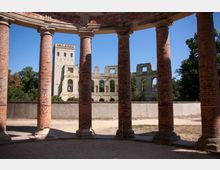 Ruine eines Backstein-Gebäudes mit offenen Wandflächen und einer Sicht auf steinerne Gebäudereste mit Fensteröffnungen, umgeben von Bäumen. Im Vordergrund stehen mehrere runde, rote Ziegel-Säulen, die zu einer kreisförmigen Konstruktion gehören.