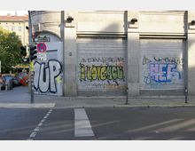 Eckgebäude mit heruntergelassenen Metallrollläden, die mit farbigen Graffiti wie "I LOVE YOU" und "UP" besprüht sind. Im Vordergrund befindet sich eine Kreuzung mit Fußgängerüberweg, Verkehrsampeln und Straßenschildern.
