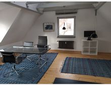 Helles Dachgeschossbüro mit schrägen Wänden, einem Glastisch und drei schwarzen Bürostühlen. Ein Fenster mit Heizkörper darunter, blaue gemusterte Teppiche auf Holzboden und ein Regal mit Monitor auf der rechten Seite.