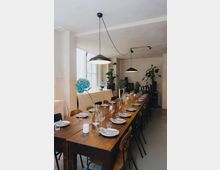 Eine lange Holztafel in einem schlichten, modernen Raum, gedeckt mit Tellern, Gläsern und Besteck. Hängelampen beleuchten den Tisch, während im Hintergrund Pflanzen und eine Anrichte mit Getränken stehen.