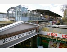Bahnhof Berlin-Zehlendorf mit überdachtem Bahnsteig und gläsernem Pavillon im oberen Bereich; im Vordergrund ein Gebäude mit der Aufschrift "Bahnhof Berlin-Zehlendorf" sowie ein Pizzastand mit der Beschriftung "La Sosta Pizza".