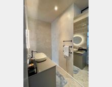 Modernes Badezimmer mit einer Waschtischkonsole aus grauem Material, darauf ein ovales Aufsatzwaschbecken und ein schwarzer Wasserhahn. An der Wand ein Handtuchhalter mit weißem Tuch, daneben ein großer Spiegel, der den Raum reflektiert; der Boden ist mit grauen Fliesen ausgelegt.