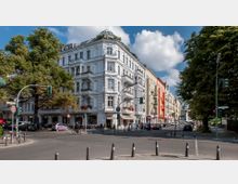 Straßenszene in einer Stadt mit einem historischen weißen Mehrfamilienhaus an einer Ecke, das mit Erkern, Balkonen und Blumen dekoriert ist. Im Hintergrund stehen bunte Gebäude, und im Vordergrund sind Fußgänger, Radfahrer, Autos sowie Ampeln an einer Kreuzung zu sehen, umgeben von Bäumen.