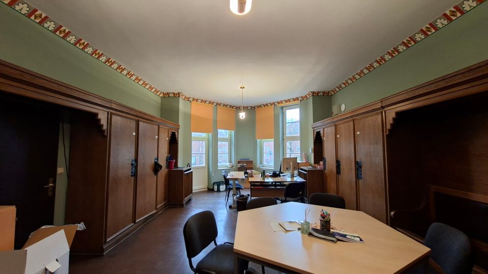 Blick in einen hohen Büro- oder Seminarraum mit grünen Wänden, umlaufender dekorativer Bordüre und großen, bogenförmig angeordneten Fenstern. Entlang der Seiten stehen dunkle Holz-Einbauten und Schränke, davor befinden sich Tische und Stühle mit Arbeitsmaterialien.